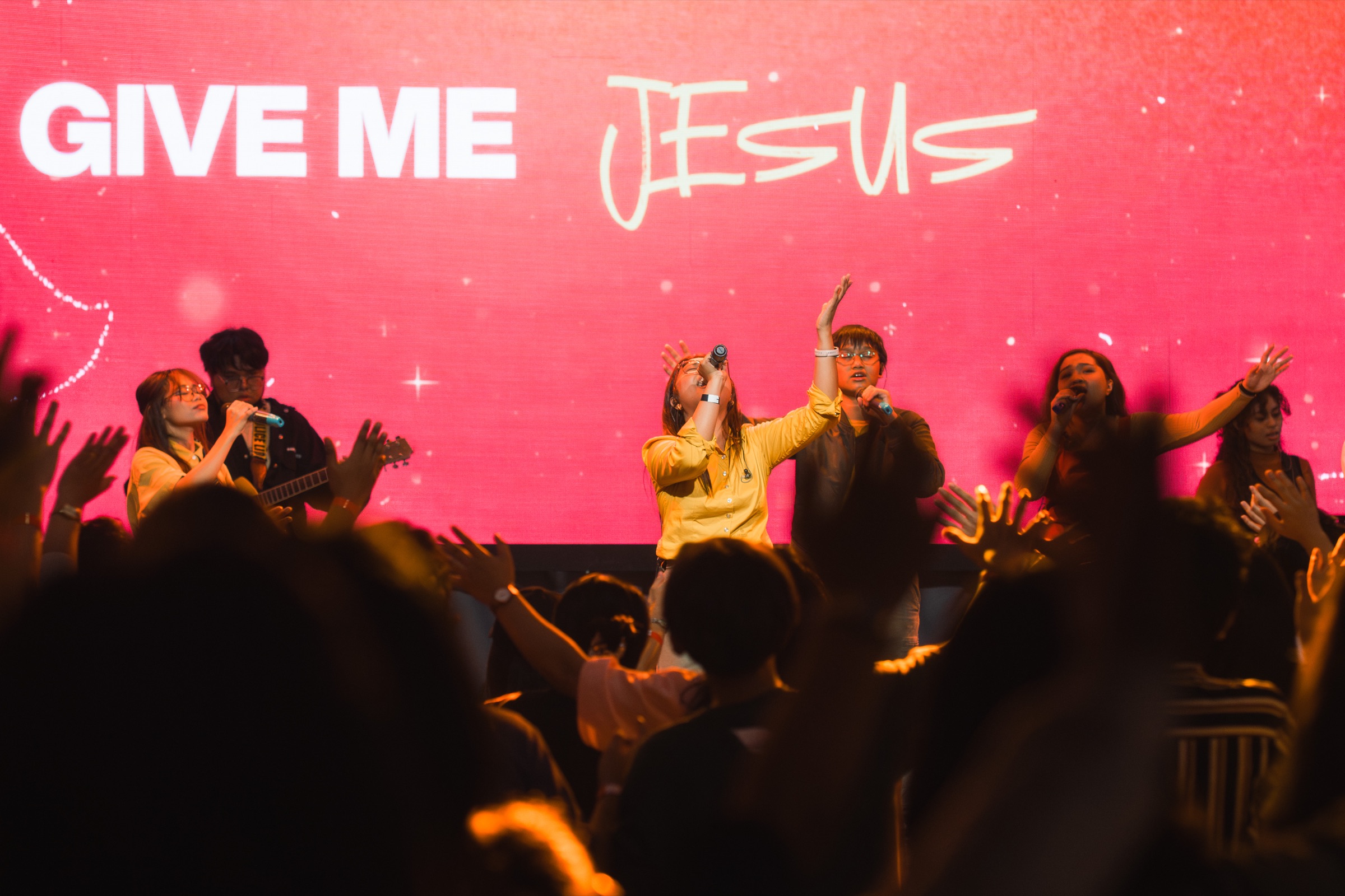 A worship team singing on stage in front of a pink "Give Me Jesus" screen as the crowd raises their hands